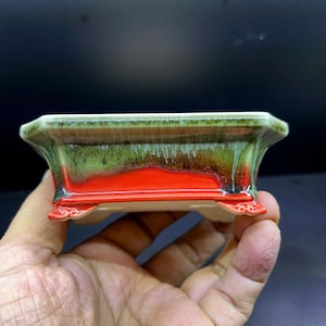 Puede incluir: Una maceta rectangular de cerámica para bonsái con una base de color rojo vibrante que se transforma en un esmalte verde intenso en el borde superior. La maceta tiene un borde acampanado, acabado brillante y cuatro patas pequeñas y curvas.
