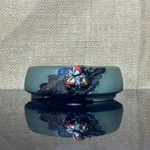 May include: A dark green ceramic bonsai pot with a black and silver glaze. The pot has a unique design with a raised, textured, black and silver design on the front. The design features a stylized, abstract shape with a red and white accent.