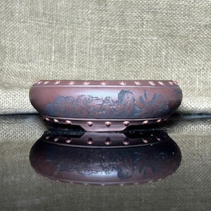 May include: A reddish-brown ceramic bonsai pot with a landscape design. The pot has a rectangular shape with rounded edges and small feet. The landscape design features mountains and trees. The pot is reflected in the surface below.