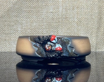May include: A brown ceramic bonsai pot with a black and white design and red accents. The pot has a textured surface and a raised rim.
