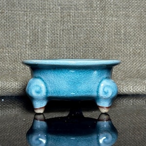 Puede incluir: Pequeña maceta de cerámica turquesa con esmalte craquelado. La maceta tiene forma rectangular con esquinas redondeadas y está sostenida por tres patas decorativas. Las patas tienen un diseño de remolino en los lados y están rematadas en marrón.