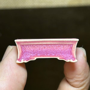 Puede incluir: Un pequeño objeto decorativo rectangular rosa y blanco. El objeto tiene un diseño curvo y adornado en la parte inferior y una parte superior plana. El color rosa tiene una apariencia texturizada. El objeto se sostiene entre dos dedos.