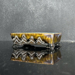 May include: A rectangular ceramic bonsai pot featuring a glossy, mottled brown and gold glaze. The exterior sides are decorated with a raised, textured relief pattern depicting a mountain range. The pot stands on four small, curved feet.