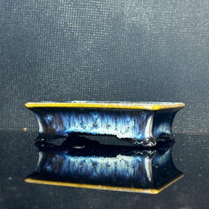 Może przedstawiać: Prostokątna ceramiczna doniczka do bonsai z reaktywnym szkliwem w odcieniach głębokiego błękitu, bieli i czerni. Krawędź wykończona jest kontrastującym musztardowym żółtym kolorem, a naczynie wspiera się na czterech małych, solidnych nóżkach.