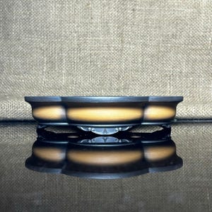 May include: A three-sectioned, ceramic bonsai pot with a dark rim and a light brown interior. The pot sits on a decorative, black, metal stand. The background is a textured, tan fabric.