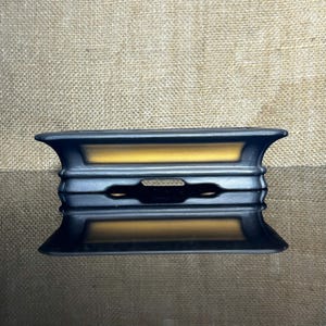 May include: A black and gold rectangular decorative object with a symmetrical design. The object has a series of horizontal ridges and a central cutout. The gold colour is visible in the centre of the object.