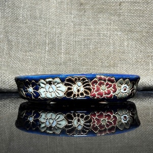 Può includere: Un piccolo piatto rotondo in ceramica blu con un motivo floreale. I fiori sono nelle tonalità del bianco, marrone, rosso e grigio. Il piatto si riflette sulla superficie sottostante.