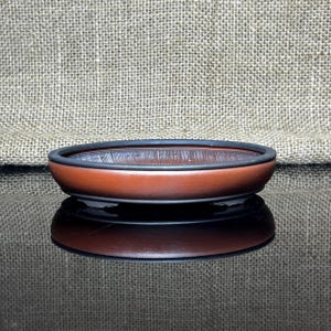 May include: A small, oval-shaped, brown ceramic bonsai pot with a black rim. The pot has a smooth, glossy finish and is reflected in the surface below. The background is a textured, tan fabric.