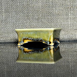 May include: A rectangular ceramic bonsai pot with a glossy, olive-green glaze. The pot has a flared shape and sits on small feet. The glaze has a textured, dripped appearance with hints of yellow and brown.