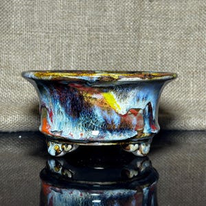 May include: A small ceramic bonsai pot with a colourful, abstract glaze. The pot has a flared rim and three small feet. The glaze includes shades of blue, orange, yellow, and brown, creating a unique, artistic appearance.