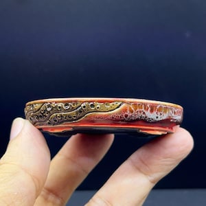 以下が含まれることがあります： ブロンズと茶色の色合いで、クレーターのような質感の縁取りが施された、浅い楕円形の陶器製盆栽鉢。本体は滑らかで光沢のある赤と白の釉薬で仕上げられています。この容器は背が低く、底には小さな脚が一体化しています。