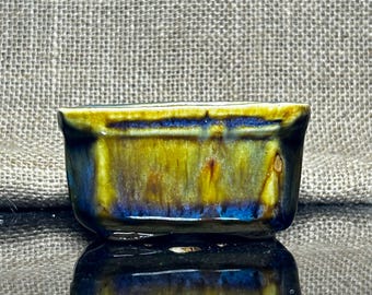 May include: A small, rectangular ceramic dish with a glossy finish. The dish features a blend of colors, including blue, yellow, and brown, creating a unique, marbled effect. The dish is likely used for serving small portions of food or as a decorative item.
