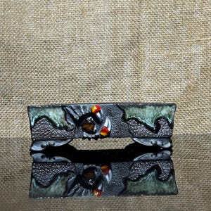 May include: A rectangular, decorative ceramic piece with a dark, textured surface. The design features claw-like elements with red, white, and yellow accents. Green, wave-like patterns adorn the edges. The piece is reflected on a glossy surface.
