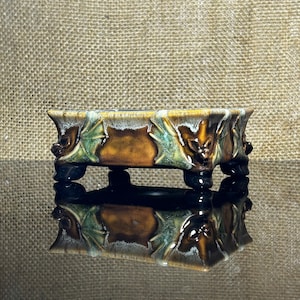 May include: A rectangular ceramic planter with a glossy brown and green glaze. The exterior features raised relief sculptures of bat faces and wings on each side. The vessel is supported by four short, dark-colored feet and has a reflective base.