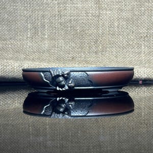 May include: A small, decorative bonsai pot with a dark red and black color scheme. The pot features a sculpted, detailed monster face on the side. The pot is sitting on a reflective surface.