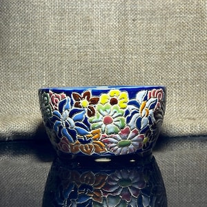 May include: A small, round ceramic bowl with a vibrant floral design. The bowl features a deep blue rim and is adorned with colorful, raised flowers in shades of pink, yellow, brown, and blue. The bowl's surface reflects the light, creating a mirrored effect.