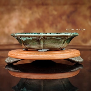 Könnte beinhalten: Ein grüner und brauner glasierter Keramik-Bonsai-Topf mit sechseckiger Form, der auf einem Terrakotta-Ständer mit drei Beinen steht. "POTTERY BY DACERA" ist in weißer Schrift auf braunem Hintergrund geschrieben.
