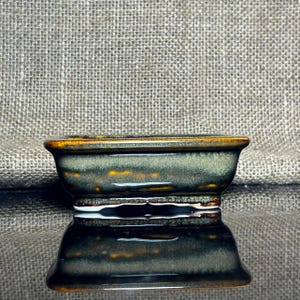 May include: A small, rectangular ceramic bonsai pot with a dark green and brown glaze. The pot has a slightly flared rim with a brown edge and sits on a small base. The pot's surface reflects the light, creating a glossy appearance.