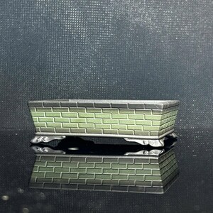 Puede incluir: Una maceta rectangular para bonsái con un patrón de ladrillo en relieve en tonos verde salvia y gris oscuro. El recipiente tiene un borde plano, lados rectos y se apoya sobre cuatro pies pequeños, curvos y ornamentados.