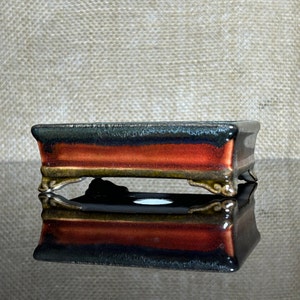 Könnte beinhalten: Ein rechteckiger Bonsai-Topf mit einer dunkelgrünen, roten und goldenen Glasur. Der Topf hat vier dekorative Füße und spiegelt sich in einer glänzenden Oberfläche.