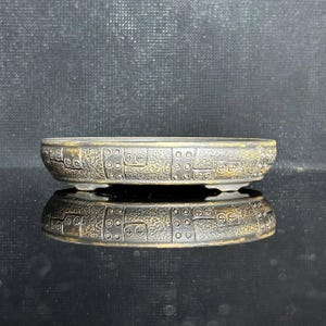 Może przedstawiać: Niska, owalna doniczka ceramiczna do bonsai o ciemnym, teksturowanym wykończeniu. Zewnętrzna strona posiada powtarzający się geometryczny wzór wypukłych kropek i segmentowanych prostokątnych bloków z subtelnym metalicznym złotym akcentem.
