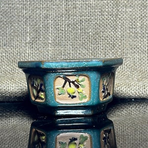 Pode incluir: Pequeno vaso de bonsai de cerâmica turquesa com formato hexagonal. O vaso apresenta painéis decorativos com um desenho floral em tons de verde, amarelo e castanho. O vaso tem um acabamento brilhante e reflete-se na superfície inferior.