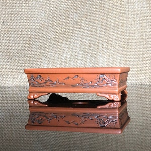 May include: A rectangular terracotta bonsai pot with a carved landscape design and four feet. The pot is brown with a glossy finish.