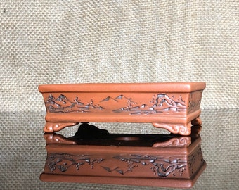 May include: A rectangular terracotta bonsai pot with a carved landscape design and four feet. The pot is brown with a glossy finish.