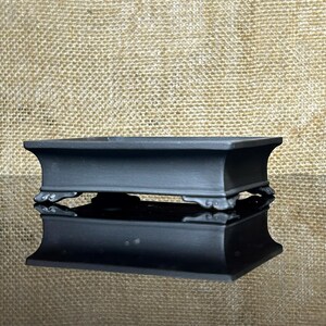 Puede incluir: Una maceta rectangular para bonsái de cerámica negra mate con bordes acampanados y cuatro pies tallados y ornamentados. El recipiente presenta un acabado oscuro y liso con una parte superior abierta y ancha, de estructura baja y estable.