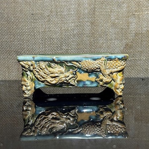 May include: A rectangular ceramic planter with a teal glaze and detailed dragon relief. The planter features a raised design of dragons and human faces on the sides and ends. The base is a dark blue colour.