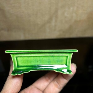 Puede incluir: Una pequeña jardinera rectangular de cerámica verde con un acabado brillante. La jardinera tiene un borde elevado y pies decorativos. El color verde vibrante y la forma única lo convierten en un artículo decorativo.