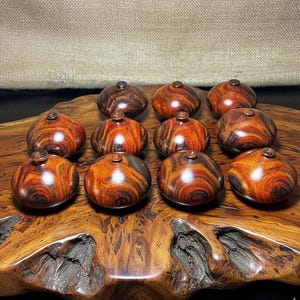 May include: A collection of twelve small, wooden decorative objects with a rounded, bulbous shape and a small, circular top. The wood grain features a striking pattern of reddish-brown and dark brown swirls. The objects are displayed on a wooden surface.