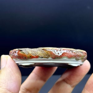 Puede incluir: Una maceta redonda y poco profunda para bonsái, hecha de cerámica con un esmalte texturizado en tonos rojo, blanco y marrón tierra. El borde presenta un acabado rugoso y moteado, mientras que la base es blanca con pequeños pies.