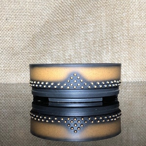 Puede incluir: Una maceta de cerámica negra con un borde marrón y puntos dorados. La maceta tiene un diseño geométrico con una forma de diamante en el centro.