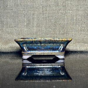 May include: A small, rectangular ceramic bonsai pot with a flared base and a rich, glossy blue glaze with hints of gold and brown. The pot's design features a raised rim and a hollowed-out base, reflecting on the dark surface below.