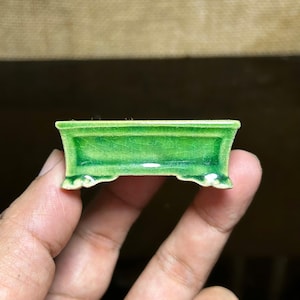 May include: A small rectangular ceramic bonsai pot featuring a vibrant emerald green glaze with a visible crackle finish. The pot has a raised rim, a recessed central panel, and four small decorative feet at the base, showcasing a glossy texture.