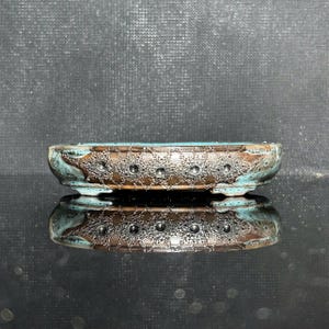 Puede incluir: Una maceta para bonsái ovalada y baja de cerámica con un exterior marrón texturizado que presenta una serie de pequeñas hendiduras circulares a lo largo del lateral. El borde y el interior muestran un esmalte turquesa vibrante.