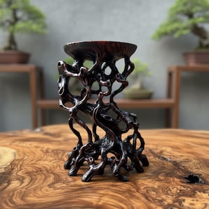 May include: A dark brown, intricately carved wooden stand with a bowl-shaped top. The stand's design resembles intertwined branches and roots, creating a natural, organic aesthetic. The stand is approximately 15 cm tall.