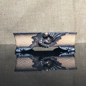 May include: A rectangular, dark brown and copper-colored ceramic bonsai pot with a raised, carved design of a flower and leaves. The pot has three legs on each side.