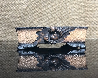 May include: A rectangular, dark brown and copper-colored ceramic bonsai pot with a raised, carved design of a flower and leaves. The pot has three legs on each side.
