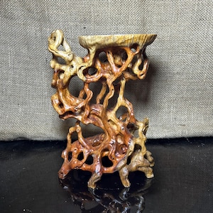 Puede incluir: Un expositor de madera hecho a mano con un diseño natural y orgánico. El soporte presenta intrincados patrones calados y un pequeño cuenco poco profundo en la parte superior. La madera tiene tonos marrones y tostados cálidos, creando una estética rústica y artística.