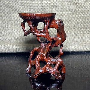 Op de afbeelding: Een handgemaakte, roodbruine houten displaystandaard met een uniek, organisch ontwerp. De standaard heeft een cirkelvormige bovenkant en een basis met een ingewikkeld, opengewerkt patroon. Het hout heeft een gepolijste afwerking, die de natuurlijke nerf en textuur benadrukt.