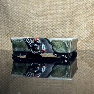 May include: A rectangular ceramic bonsai pot featuring a mottled green and grey glaze with a textured black relief sculpture of a crab on the front. The vessel is supported by four short, dark, curved feet and has a smooth, glossy finish.