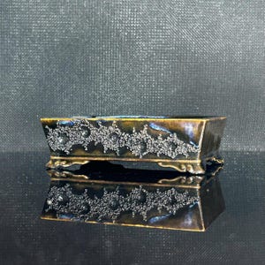 May include: A rectangular ceramic bonsai pot featuring a dark brown and metallic blue glazed finish. The exterior is decorated with a raised, textured, crystalline pattern along the sides. The pot stands on four small, ornate, curved feet.
