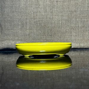 Puede incluir: Un pequeño plato ovalado de cerámica amarillo brillante. El plato tiene un acabado brillante y se refleja en la superficie inferior. El fondo es una tela gris texturizada.