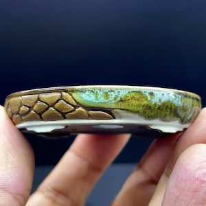 Könnte beinhalten: Eine flache Bonsaischale aus Keramik mit einer strukturierten braunen Außenseite, die auf einer Seite ein geometrisches Schuppenmuster aufweist, und einer glatten, glänzenden grün-weißen Glasur auf der anderen Seite. Das Gefäß hat kleine Füße.