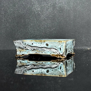 Puede incluir: Una maceta rectangular de cerámica para bonsái con un esmalte reactivo en tonos azul claro, blanco y marrón oscuro. La maceta tiene una superficie texturizada y moteada, un borde ancho y se apoya sobre cuatro pies pequeños y ornamentados.