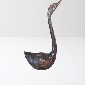 Può includere: Una statuetta di cigno color marrone scuro con una texture particolare, un collo lungo e curvo e un becco allungato. La superficie presenta un aspetto maculato e usurato con sottili sfumature nere e ramate su tutto il corpo e il collo.