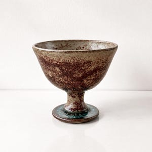 Può includere: Un calice in ceramica fatto a mano con una coppa ampia e una base a piedistallo. Il calice ha una combinazione di colori marrone e beige screziato con un tocco di verde acqua alla base. Il calice viene probabilmente utilizzato per servire bevande.