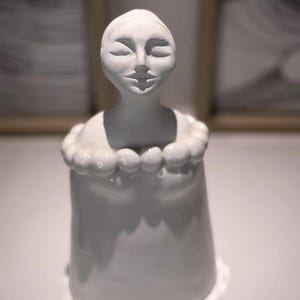 May include: A matte white ceramic sculpture of a stylized figure with a smooth, rounded head and closed eyes. The figure features a prominent collar made of a series of uniform, spherical beads encircling the neck, resting atop a cylindrical base.